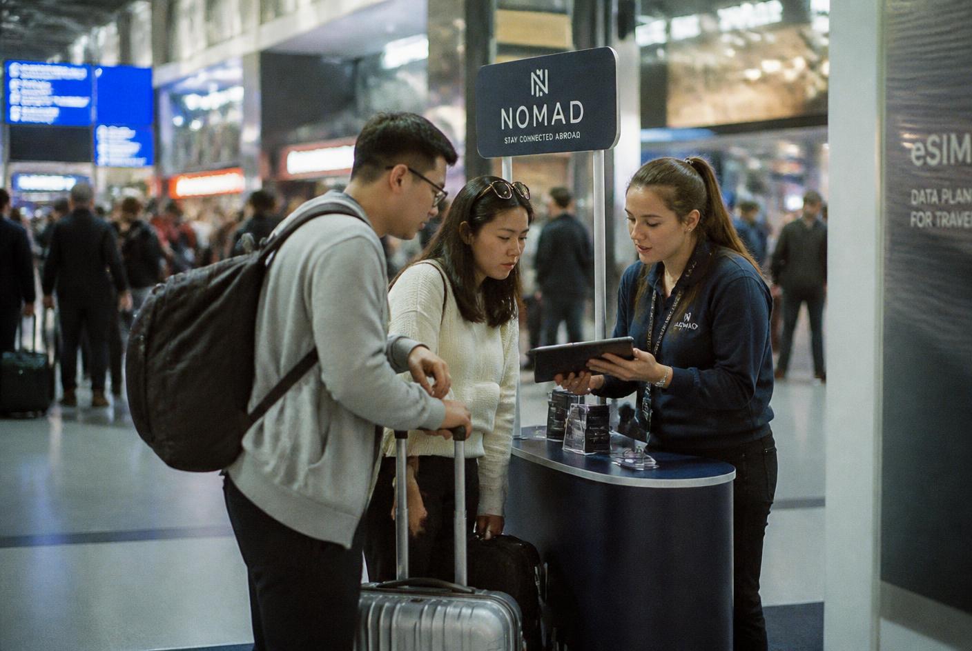 Sales agent showing a tablet to two travellers at a promotion desk in an airport concourse